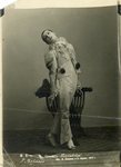 Photograph of Samuil Vermel as Pierrot, his costume white with black pompoms, standing, head back, with a hand on a chair.