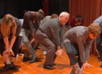 Community members of diverse ages, races, ethnicities, and genders rehearse a gesture on stage. Each stands in a loose fifth position with right foot in front, bent at the knees in a plié, and bent at the waist as they bring their wrists together in front of their bent knees, hands splayed outward, as if in the shape of a bird or flower. All look down at their feet except an older gentleman at center, who looks up at the leader of the group outside of the frame.