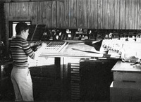 Black-and-white photo. Cohen faces the row of type cabinets, holding a composing stick with type to redistribute. Type drawer is set on the angled bank on top of the cabinets.