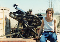 In jeans and a tank top, Cohen sits on a flatbed truck with her back to crates and a loaded press with a puzzled and somewhat resigned expression. Farm across the road in the background.