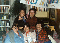 Color photo in Cohen’s living room. The group is dressed festively with bandanas, scarves, and ties, with Hamilton in a mustache and funny glasses. Feminist library in the background.
