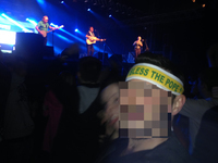 A man in a concert crowd wears a headband reading “God Bless the Pope