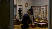 A man kneels at a chabudai in front of room screen divider with black calligraphy printed on it.
