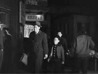 A man and boy walk at night beside lit store signs with black calligraphy, in black and white cinematography.