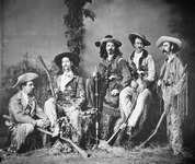 A group of male actors in elaborate American West-style costumes poses for the camera. The men wear hats and fringe trimmed buckskin coats. The men hold rifles and wear hats, kerchiefs, and belts with large buckles. Grass and plants on studio floor with a backdrop in background. Typed caption on the mat, “Furman Rochester.”