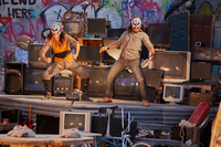 Two actors wearing white and colored masks stand slightly crouching on top of a platform with defunct televisions surrounding them. Graffiti covers the walls behind.