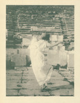 Photograph of Isadora Duncan dancing among ancient ruins in Athens. She wears a white chiton (a Greek-styled draping dress) and sandals, posing with arms outward and facing upward, mid-step with one leg bent.
