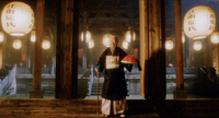 An older man with a cane in one hand and hat in the other stands in front of a courtyard and pillars. Calligraphy is visible on the many lanterns.