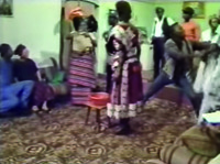 Film still frame of a large gathering in room with patterned carpet and sofas