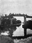 A black-and-white photo showing a bridge over a canal, with more than a dozen figures on the bridge. The shadows of the bridge and the people are reflected in the water below the bridge.