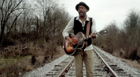 Figure 12. Darius Rucker portrays the persona of an itinerant country musician by playing and singing while walking on railroad tracks, wearing contemporary clothing that looks older, and playing a Gibson archtop style acoustic guitar.