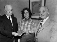 1970s era photograph of a younger woman wearing blue jeans and a plaid shirt stands between two older men wearing suits; the man to her left holds out a small book, and she accepts it.