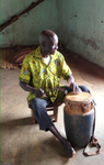 A man wearing an African print shirt is seated playing a drum with a curved stick. He looks out the doorway to his left into the light.