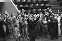 Bows at the end of the performance suggest the participants’ sense of accomplishment. About thirty cast members stand at the end of a an entrance leading to the arena floor with basketball arena seats visible behind them. Instead of bowing, they applaud one another and their audience. Some applaud Jones himself, who looks out at the audience with arms stretched overhead, waving with a serious expression on his face.