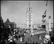 A black-and-white photograph of the white campanile tower at the center of the Dreamland amusement park on Coney Island. To its right is an imitation of the Doge’s Palace. An extended boardwalk in the foreground is filled with amusement park patrons.