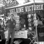 In jeans and a plaid flannel shirt, Cohen stands by the HV display. “Helaine Victoria Women of History” banner hangs above the table. Shoppers in background.