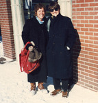 Color photo. The two stand outside a brick building in long wool coats and boots, smiling on a sunny, snowy day. Zolot wears sunglasses. Cohen squints from the glare.