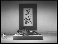 A naked man and woman lie in a studio in front of a giant posterboard with black calligraphy on white on a black matte background, in black and white cinematography.