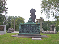 Color photograph of monuments at Homewood Cemetery, Pittsburgh, of various sizes and styles. In front is a bronze sarcophagus monument, with angels holding wreaths on the corners, and draped laurels in the front panels. Atop the monument is a draped urn. Behind the sarcophagus monument is a tall tree stump monument with names in the place of missing limbs. Other monuments behind these include ledger slabs with headstones in different styles in granite and marble and a tall neoclassical monument with Corinthian columns on each of the four corners.