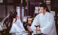 Wei Wuxian (left) and Lan Wangji (right) in a study at Cloud Recesses. They are both clad in white robes and have long hair. Lan looks intently at a jar of Chinese brushes as he chooses one, while Wei looks on with one arm supporting his head.