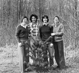 Four women stand behind a small pine tree, one in a plaid flannel shirt, others in sweaters. Trees are in the background in a winter silhouette. All women smile toward the viewer.