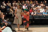 A smiling woman is escorted by a man who is kneeling. He wears knee pads and a green headpiece. They walk in an indoor theater, with audiences on two sides.
