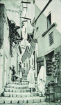 Black-and-white photograph of a narrow stepped street with people on it, peering into shops and talking to one another.