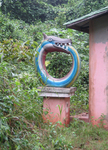 A sculpture of a snake eating its tail, painted like a rainbow, next to a small house in a forested setting.