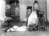 Black-and-white photograph of a woman in a wedding dress lying on the floor, presumably dead, while another actor looks at her