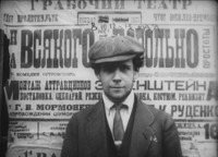In this film still, Eisenstein, in blazer, tie, and cap, looks out at the viewer of the film. The background behind him is filled with a large poster that introduces the term "montage of attractions" and advertises the May 8, 10, 12, and 13 performances of Wiseman.