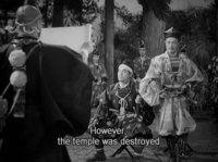 A man reads from an unfurled scroll to a seated warrior and his standing companions. White roman subtitles are at the bottom.