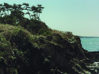 Landscape shot of a rocky bluff overlooking the ocean, sparsely dotted with shrubs and greenery as well as trees at its top. From this extreme distance, Aochi is barely visible in his grey tweeds as he stands on an unseen path going up the cliff.