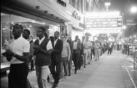 In 1963, a group of African American and white protesters march down a Raleigh sidewalk at night, clapping and singing freedom songs.