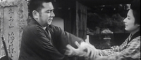 A man grabs a woman's wrists in front of a gate. Calligraphy is visible on the stone behind them.