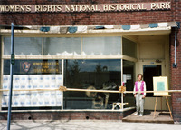 Cohen stands in front of Suffrage Press print shop. Press visible in window; park name above; gold ribbon across. Next window says “Women's Education and Culture Center.”