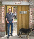 A photo of a man standing at a wooden doorway, alongside a vertical nameplate with calligraphic text.
