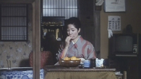 A woman sits and eats from a bowl on a low table. Calligraphy is visible on signs and posters in the background.