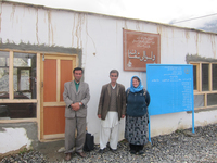 Author standing with deputy governor of Shughnan district and his deputy. Author is wearing blue hijab and long traditional dress. Deputy governor has shalwar kameez with suit coat over it. The two men and the author are standing in front of the governor’s office, which is still being constructed. The title of the building is Dari in the background.