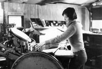 Black-and-white side view of Poore feeding a piece of paper into the press. She wears a sweater, a shirt, and jeans.