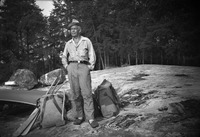 A black-and-white portrait of Olson standing on the shore next to a pack.