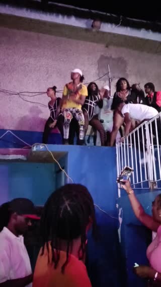 Video recorded during the night at a street dance party. Around 12 dancers danced on a balcony or a roof. 3 females and 9 males. They are performing the same choreography. The same dancehall moves of a set choreography of the songs played.