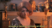 Screen capture of Dorian Corey in full makeup and wig, sitting before trophies on a mantle. The caption reads “reading is the real artform of insult.”