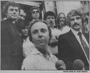 Image of a young woman in front of microphones, surrounded by people, all looking toward the viewer.