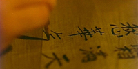 Close-up image of black calligraphy being written in pencil on textured wood.