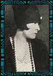Side portrait. Johnson wears a simple cloche hat almost covering her eyes. She wears a dark jacket, a V-neck knit top, and a long string of beads.
