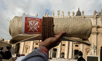 Color photograph of a rolled-up, woven Tongan mat being held aloft. In the background is the facade of St. Peter’s Basilica in Rome.