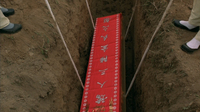 A coffin is lowered into a dug pit with a banner with white calligraphy printed on it.