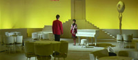 A long shot of the Club Alulu, with walls, furniture, stage settings and statuary all bathed in a uniform yellow lighting. In the background center is Chiharu next to a white piano, being approached by Ōtsuka in a red dinner jacket. Behind and above Chiharu is a set of stairs leading up to a door; the yellow light obscures depth and conceals the fact that the door and stairs are in fact freestanding and unattached to the wall.