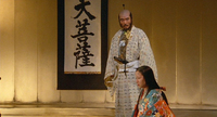 A man stands in front of a kneeling woman with a banner with black calligraphy hanging on the wall behind him.