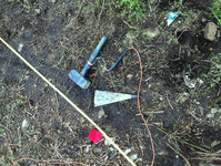 Mason’s hammer, steel plate, measuring tape, and red flag are shown in the photograph laying on the ground.